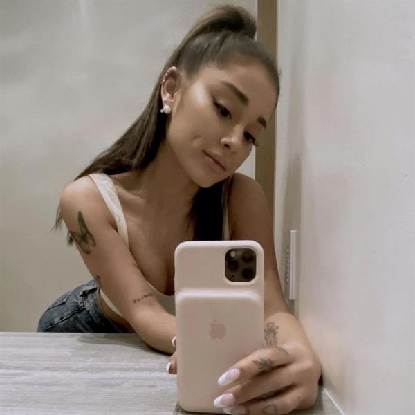 Ariana Grande boobs showing nice cleavage with her tits in a sexy little white tank top leaning forward on the counter for a selfie.